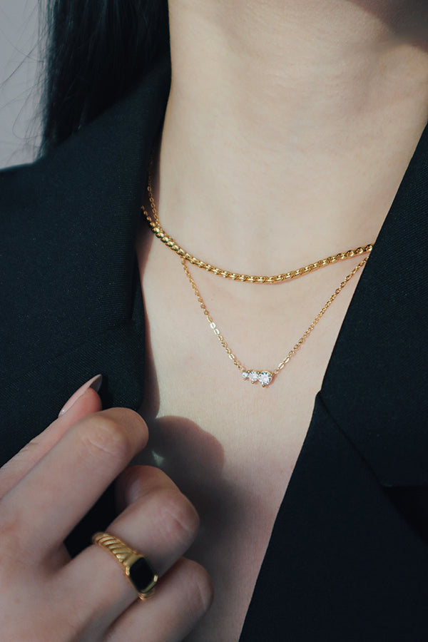 A person wearing two layered gold necklaces: a top textured chain and a lower delicate chain with a cluster of three clear cubic zirconia stones, styled with workwear.