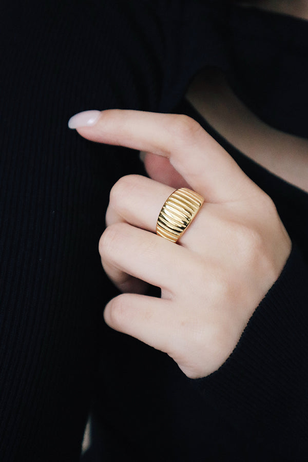 A chunky gold ring with a ribbed or fluted design worn on a finger, featuring a polished finish.