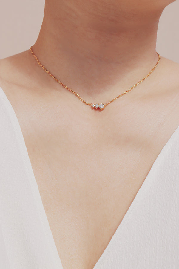 A delicate gold necklace with a horizontal cluster of three clear cubic zirconia stones, worn on a person's neck.