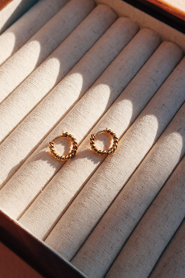 一對金色扭紋耳環擺放在米色首飾盤上,在溫暖的陽光照射下呈現出精緻的扭轉設計。
