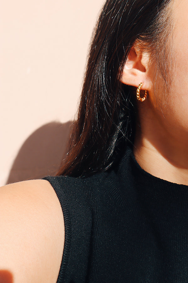 一位女性佩戴扭紋金色圈圈耳環的側面特寫,搭配無袖黑色上衣,背景為柔和的粉紅色。