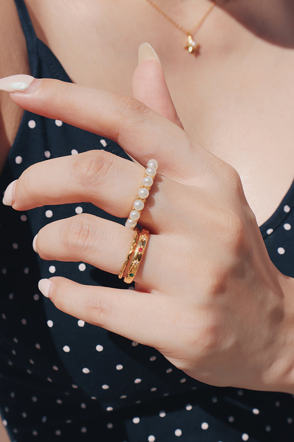 A close-up of a female hand with light-colored manicured nails, wearing a gold ring with a line of small white pearls on the middle finger and a textured gold ring on the ring finger. The woman is wearing a dark top with white polka dots, and a delicate gold necklace is visible around her neck.