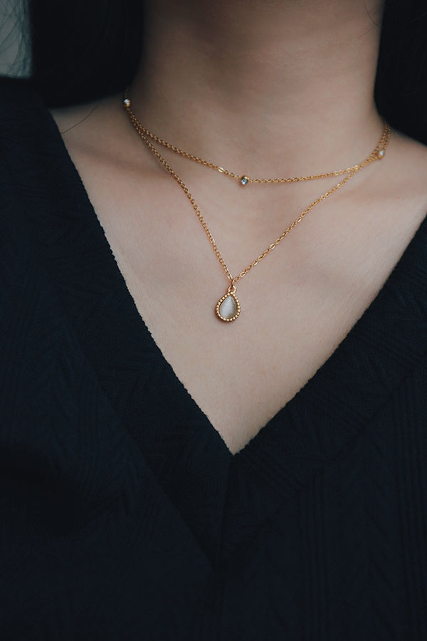 Layered 18K gold-plated necklace set with a teardrop-shaped pendant showcasing a mother-of-pearl inlay, paired with a dainty chain adorned with small cubic zirconia stones, styled with a black V-neck top.