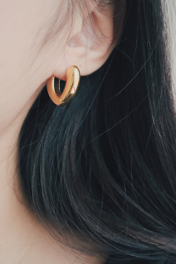 A close-up image showcasing a pair of 18K gold-plated heart-shaped hoop earrings on a womans ear. The earrings have a bold and modern design with a curved shape that resembles a heart, giving them a trendy and luxurious appeal.