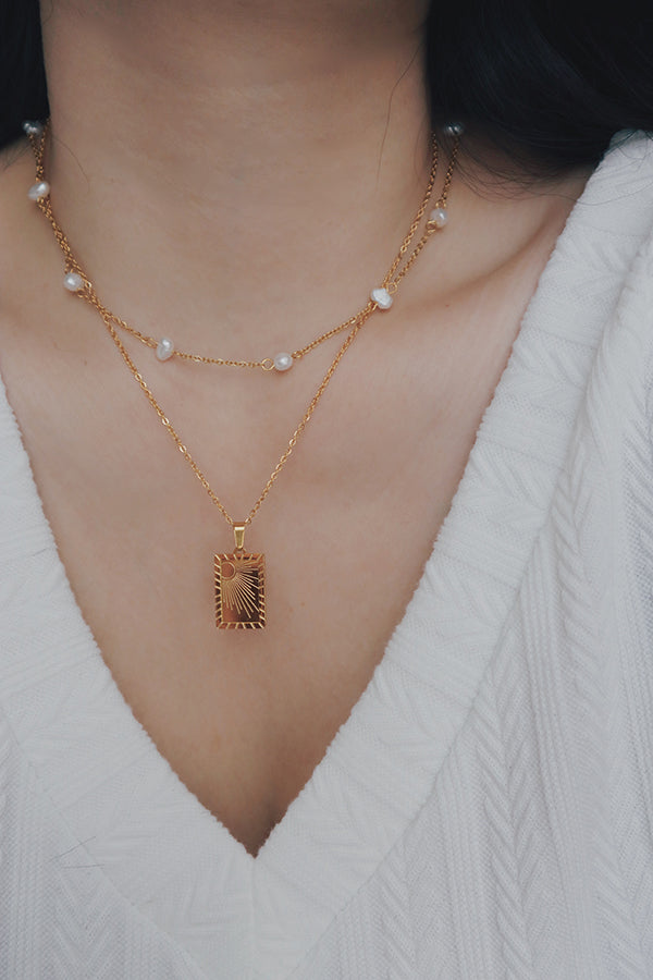 Close-up of a woman wearing an 18K gold-plated Savita Sun necklace, featuring a rectangular pendant with a sunburst engraving, styled with a delicate pearl choker and a white textured V-neck top.