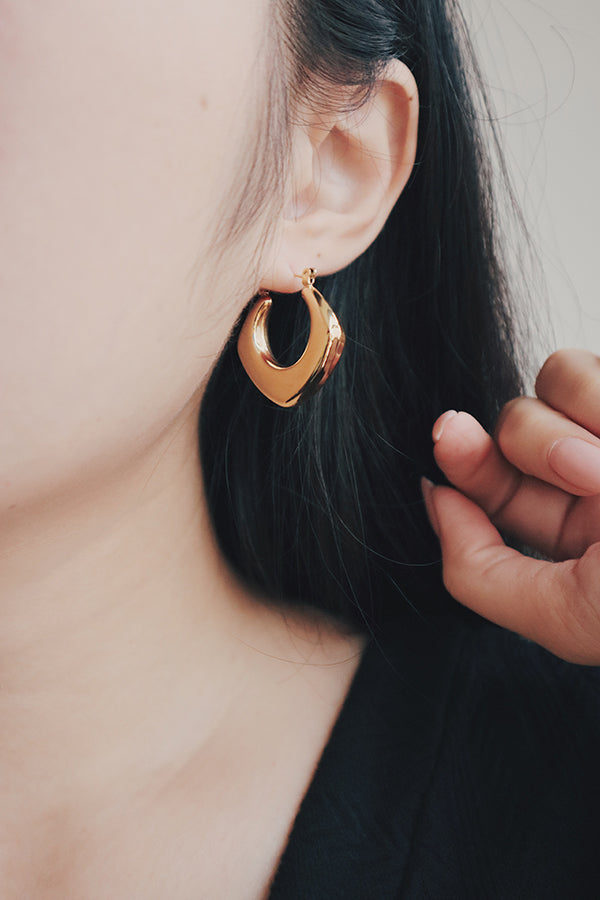 A close-up image of a womans ear wearing a gold square hoop earring with a smooth, polished finish. The earring features a bold, curved design with a geometric shape, adding a modern and sophisticated touch to her look. The shiny surface of the earring catches the light, highlighting its luxurious 18K gold plating.