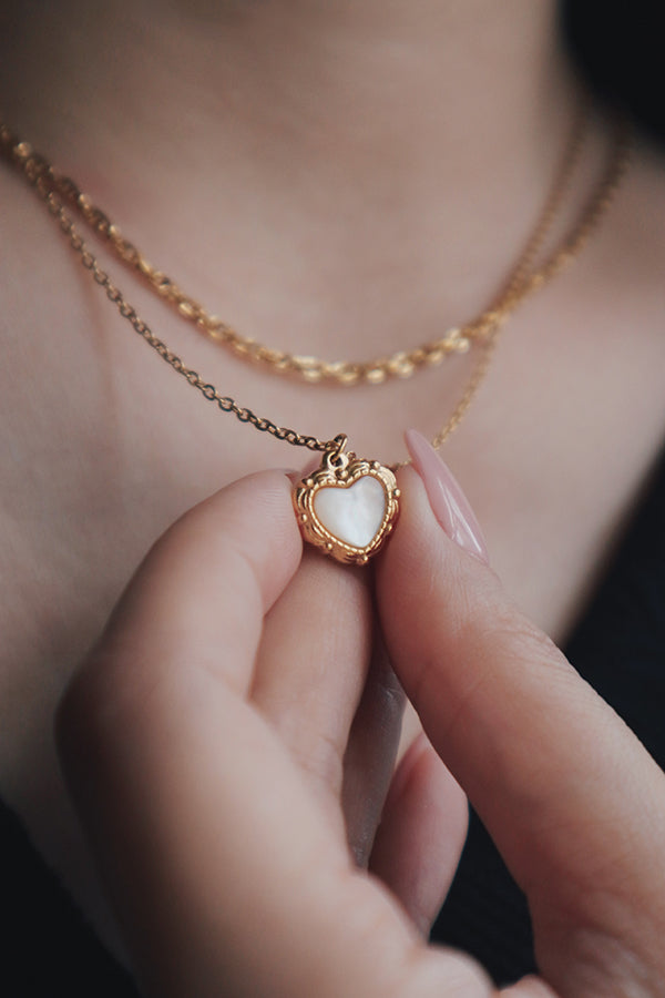 Close-up of a hand holding a heart-shaped pendant with a mother-of-pearl inlay, showcasing the detailed gold beaded frame. The necklace is layered with a second gold chain and worn with a black top.
