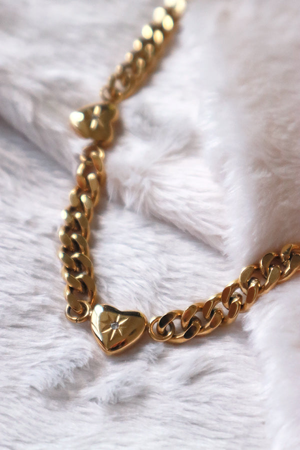 一條奢華的18K鍍金頸鍊,採用古巴鍊設計,鍊條上間隔鑲嵌著多個心形墜飾,每個心形中央都點綴著一顆微小的閃亮寶石,質感高雅。