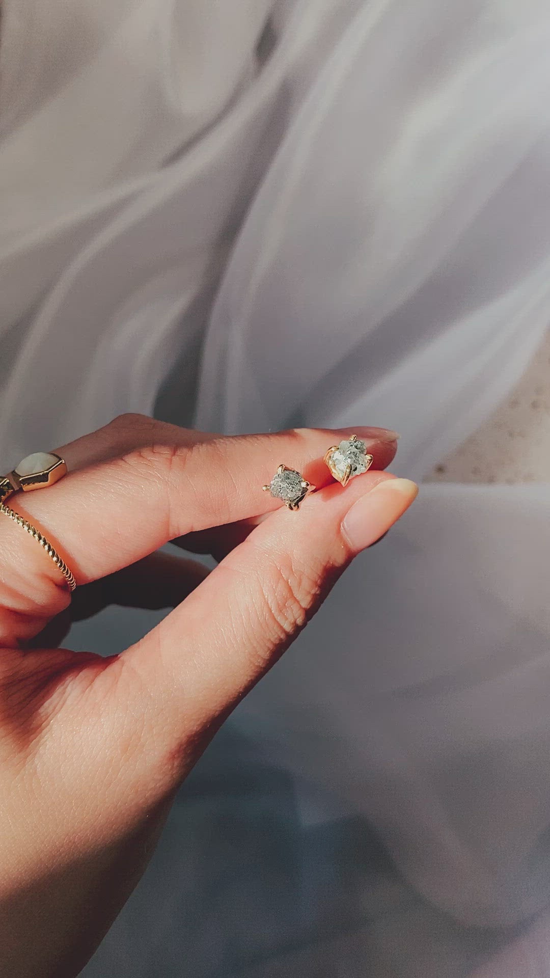 A short video clip showing a female hand, with gold rings on her fingers, holding two small gold stud earrings, each with a sparkling clear, raw diamond or crystal. The hand gently moves, displaying the earrings against a softly draped white translucent fabric.