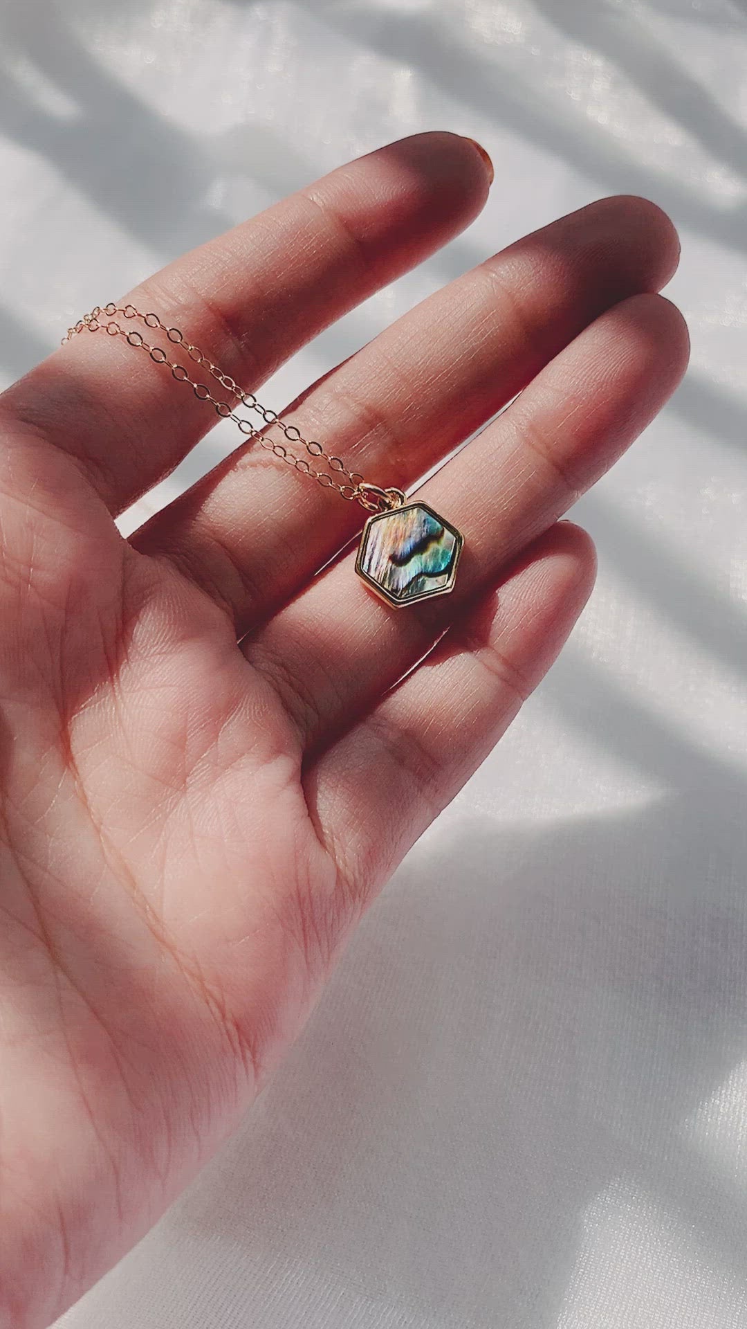 A short video clip showing a female hand holding a delicate gold chain necklace with a hexagonal abalone shell pendant. The hand gently turns the pendant, revealing its iridescent blue, green, and purple hues under sunlight. The background is a white textured fabric with shadows.
