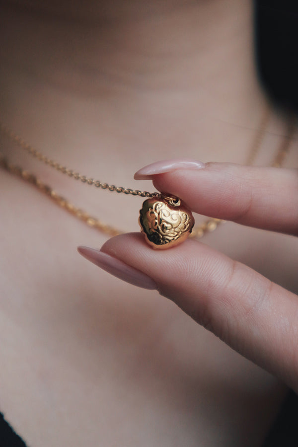 Close-up of a hand holding the back side of a heart-shaped gold pendant with intricate engravings, highlighting the design details. The necklace is layered with a second chain.