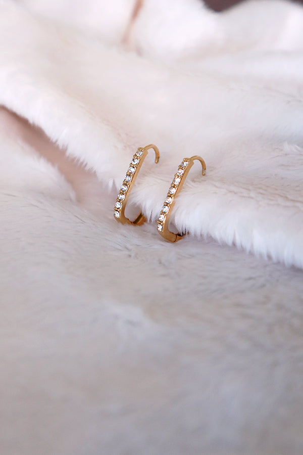 An overhead shot of a pair of gold rectangular hoop earrings, each embellished with a line of sparkling clear cubic zirconia stones. They are resting on a soft, fluffy white surface.
