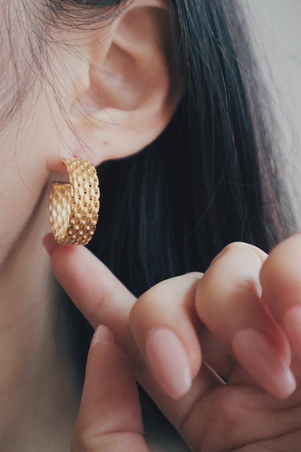 A detailed shot of a woman wearing textured gold hoop earrings. The earrings have a wide band with a woven texture, reflecting light to create a luxurious and eye-catching look, perfect for summer styling.