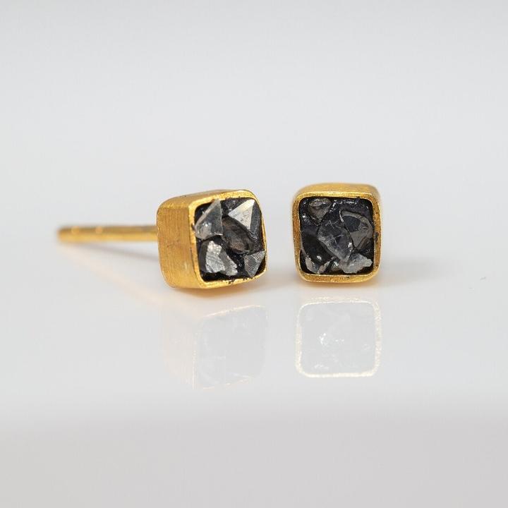A close-up, eye-level shot of a pair of gold-toned square stud earrings, each featuring a raw, crushed black diamond or stone inlay. The earrings are displayed on a white reflective surface.