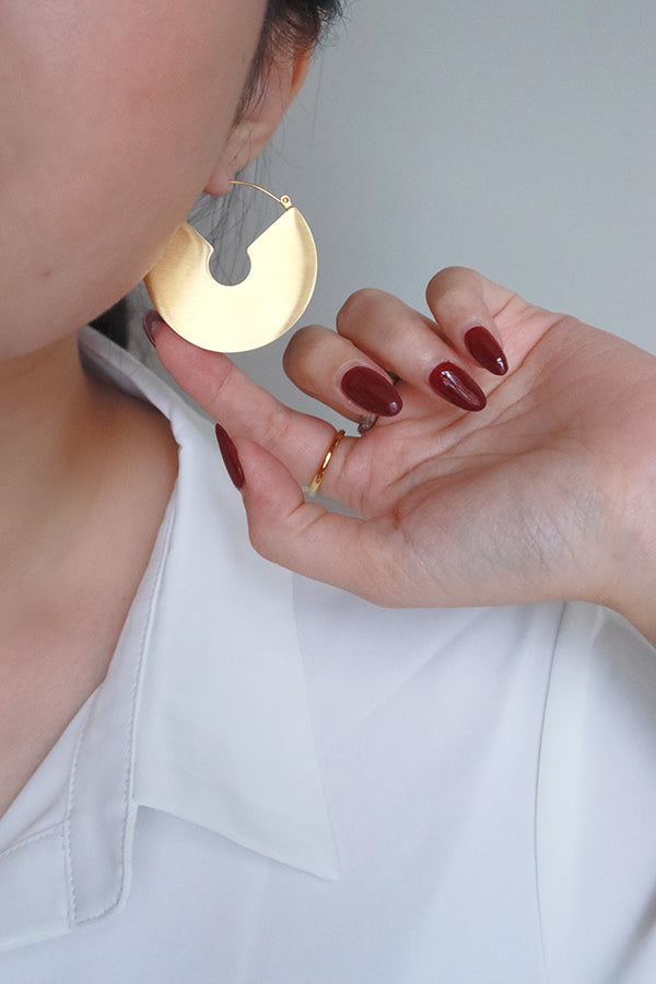 一隻耳朵佩戴著一款大型圓形扁平鍍金耳環,表面呈現啞光拉絲質感,中央帶有獨特的弧形切口設計,整體風格散發著波西米亞風情。