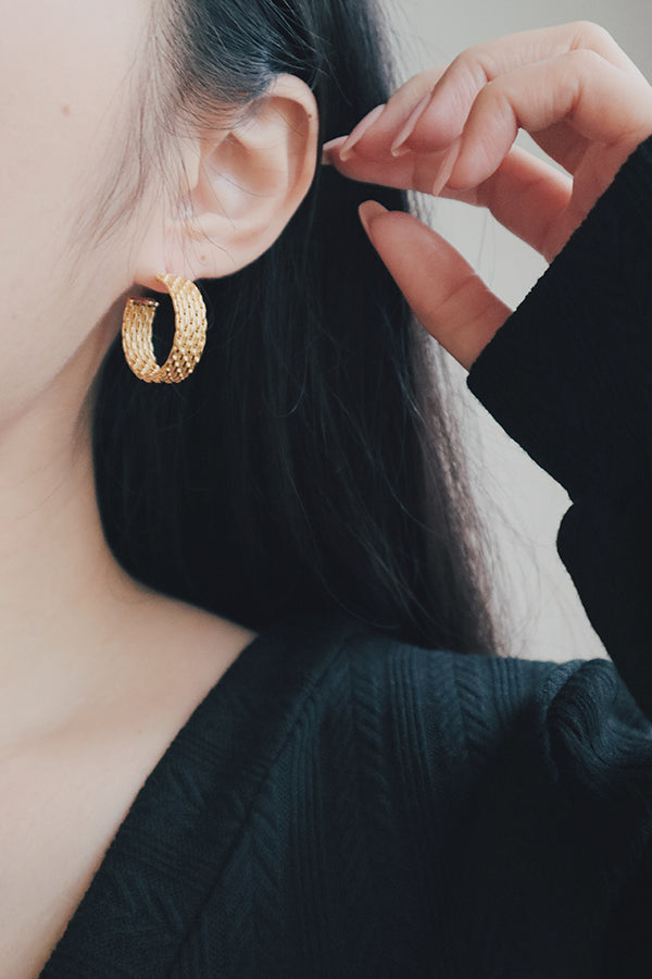 A close-up image of a woman's ear wearing a bold, textured gold-plated hoop earring. The earring features a detailed, raised pattern that adds depth and dimension, creating a striking and elegant accessory.