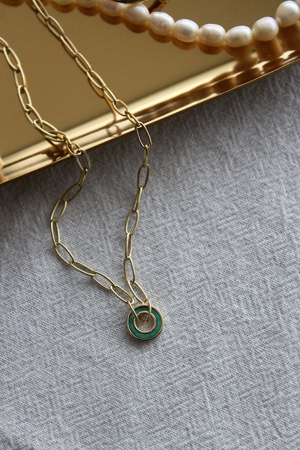 A gold paperclip chain necklace with a small circular green malachite pendant, laid on a light gray textured fabric. In the background, a gold tray and a string of pearls are visible.