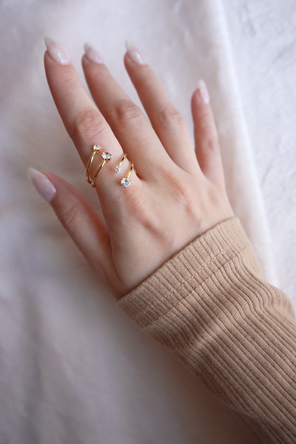 A person's hand wearing a modern gold ring with a layered wrap design, adorned with three clear cubic zirconia stones.