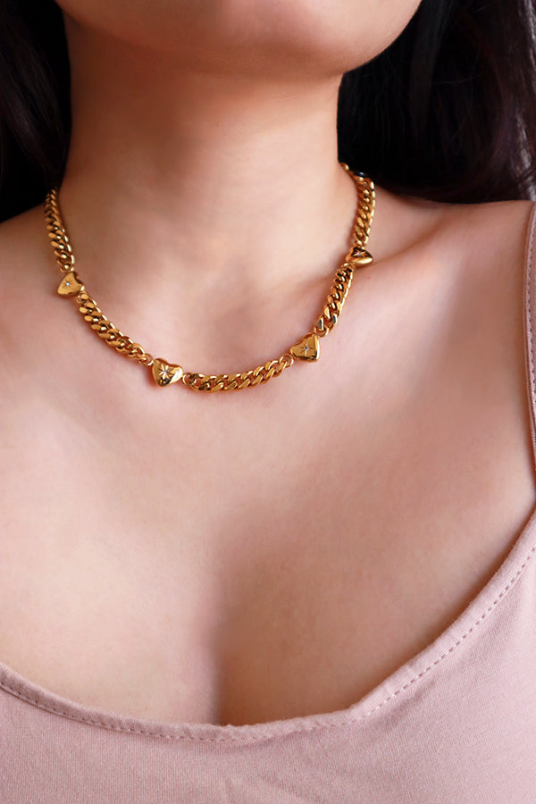 一位女性的頸部佩戴著一條散發可愛氛圍的金色古巴鍊項鍊,粗獷的鍊條與其間隔的心形墜飾形成鮮明對比,時尚又甜美。