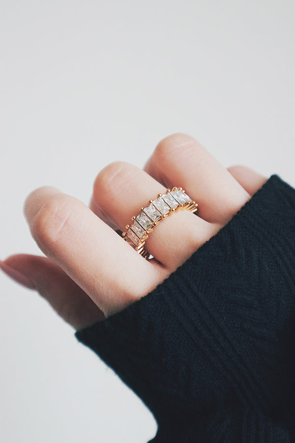 A hand wrapped in a cozy black knit sleeve showcases a stunning gold eternity ring adorned with rectangular-cut cubic zirconia stones. The ring's thick band and sparkling stones create a powerful statement piece that radiates luxury and style. The hand's soft, natural lighting enhances the ring's brilliance, making it the focal point of the image.