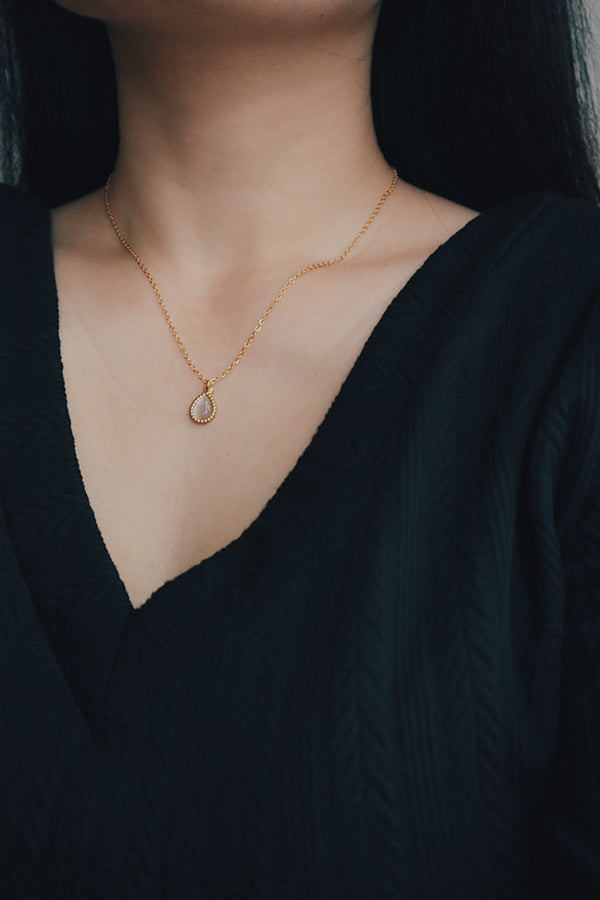 Close-up of a woman wearing a dainty gold-plated necklace with a teardrop-shaped pendant featuring a cat's eye gemstone, styled with a black V-neck top.
