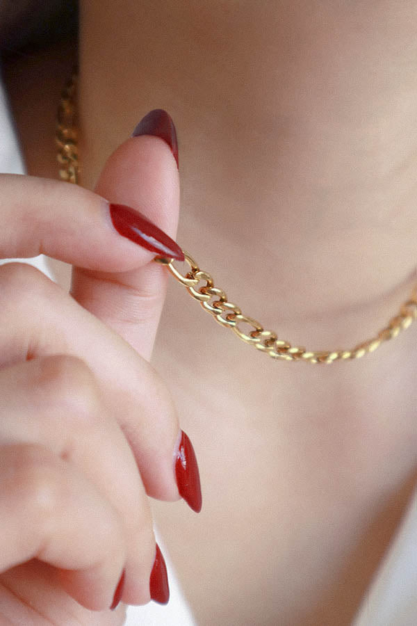 A close-up of a female's hand with long, dark red manicured nails, delicately holding a segment of a shiny gold Figaro chain choker near her neck, emphasizing the details of the links. A portion of a white collared shirt is visible.