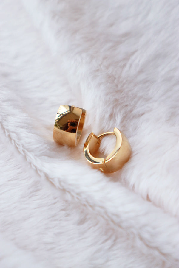 A pair of shiny, wide gold hoop earrings with a polished, rounded surface, resting on a soft white textured surface.