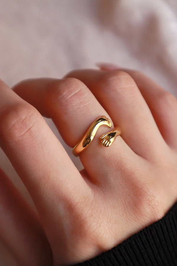 A close-up of a woman's finger wearing a polished gold "hug" ring, showing the intricate details of the small hands embracing.