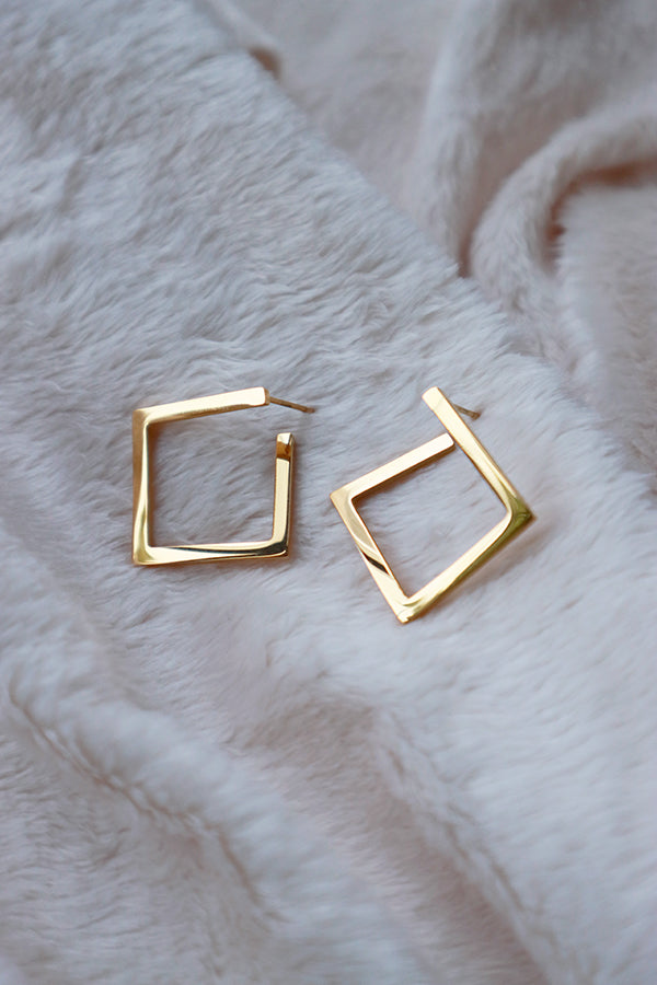 A pair of simple, polished gold square hoop earrings, lying on a soft white textured surface, showcasing their modern design.