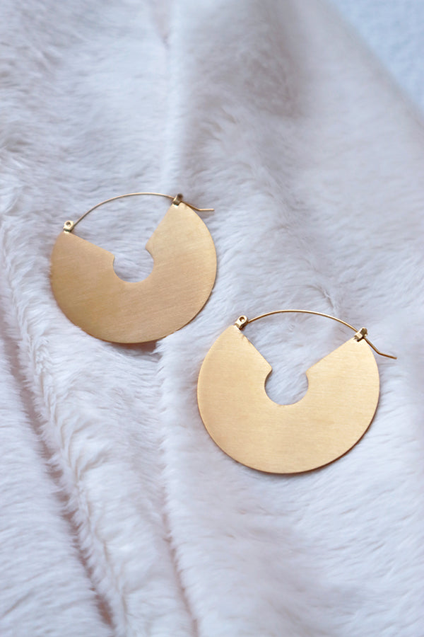 A pair of large, flat, matte gold-colored stainless steel hoop earrings with a distinctive inner cutout, lying on a soft white textured surface.