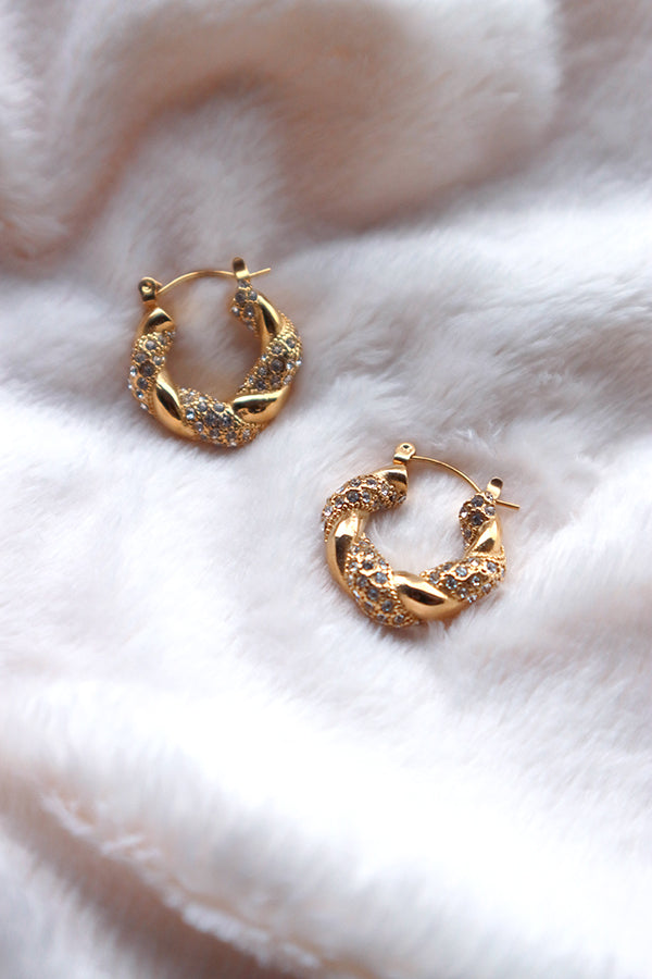 A pair of stainless steel gold-plated twisted hoop earrings, adorned with numerous cubic zirconia stones, resting on a soft white textured surface.