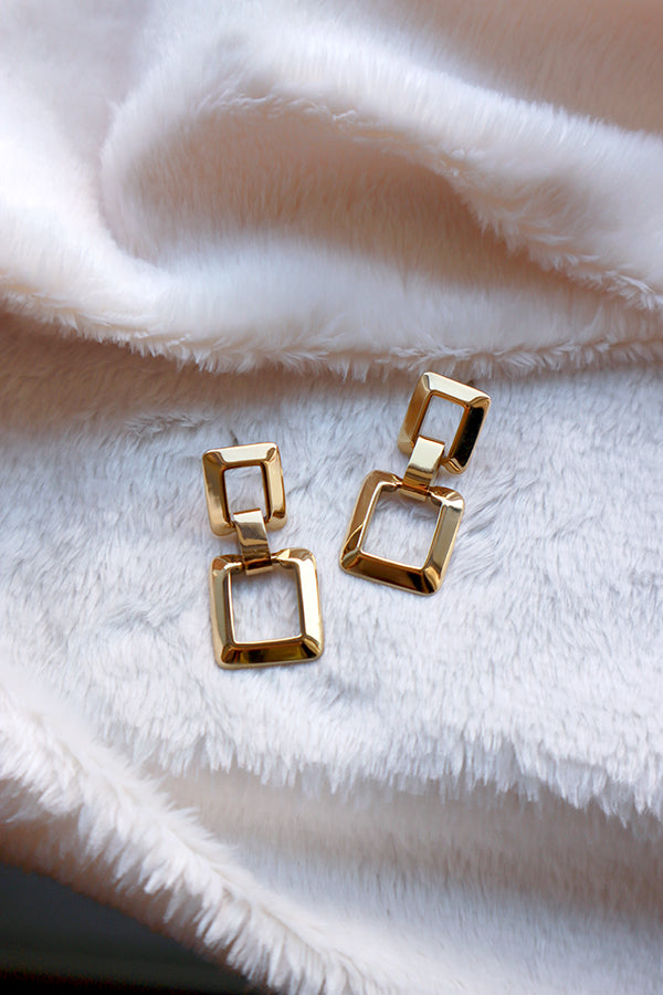 A pair of shiny stainless steel gold-colored square linked earrings, lying on a soft white textured surface, showcasing their geometric design.