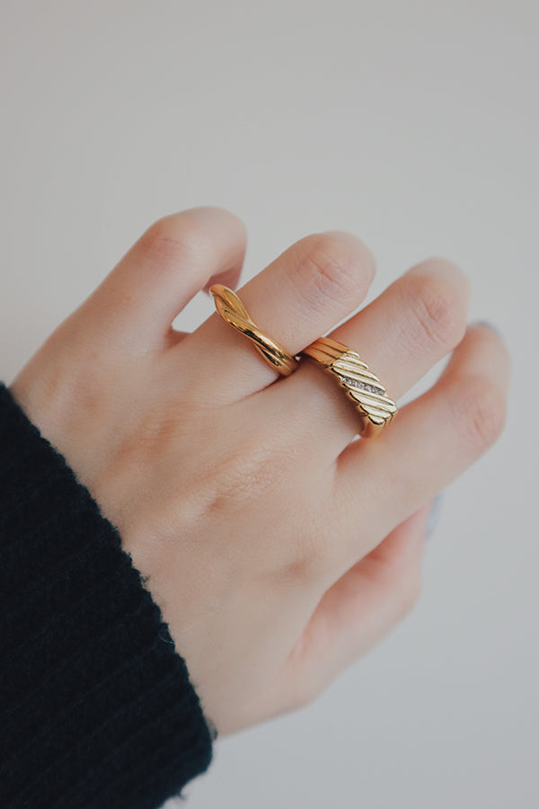 一隻女性手部特寫,無名指上戴著兩枚金戒指:一枚是波浪形的雕塑感戒指,另一枚是較厚的戒指,帶有紋理條紋設計,並鑲嵌著一排斜向的小閃亮透明寶石。這位女性穿著一件深色長袖服裝。