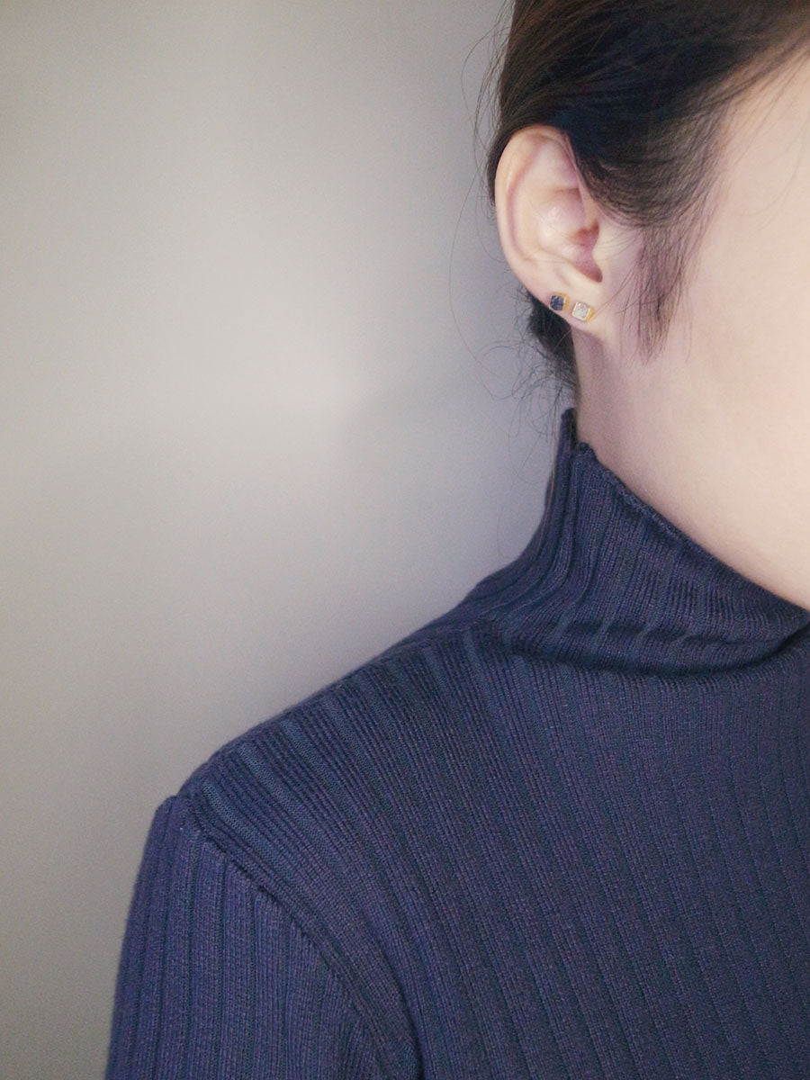 A close-up side profile of a woman's ear and neck, showcasing two small gold-toned square stud earrings. One features a crushed dark stone, and the other a crushed light stone. The woman has dark hair and is wearing a dark blue ribbed turtleneck sweater.