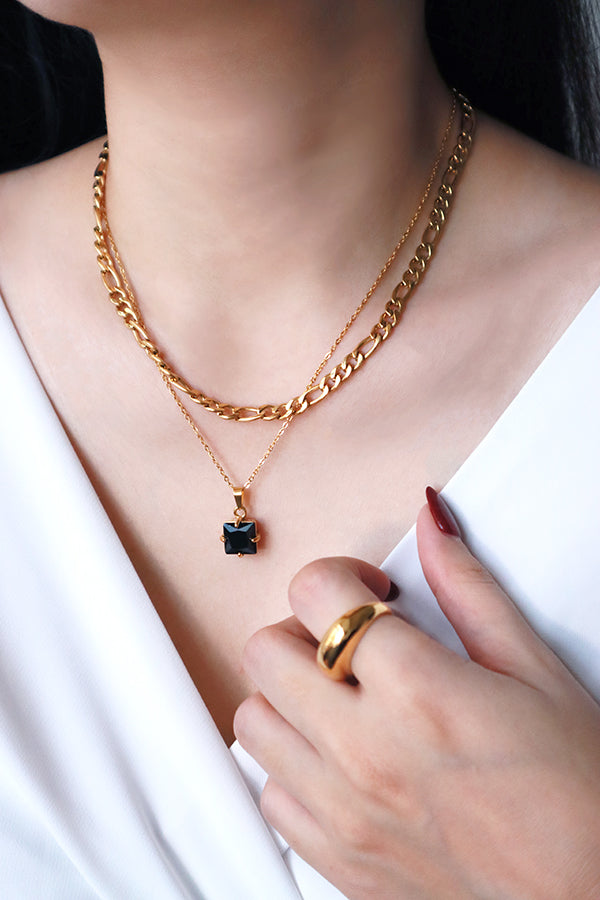 一位女性胸部和肩部的特寫,展示兩條疊戴的金色項鍊。上面一條是帶有珠飾的粗費加洛鏈條,下面一條是帶有方形切割黑色水鑽吊墜的精緻鏈條。這位女性穿著白色上衣,無名指上戴著一枚金戒指,並留著深紅色美甲。