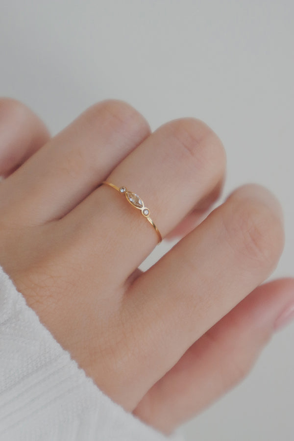 A delicate gold ring featuring a marquise-shaped clear cubic zirconia stone flanked by two smaller round stones, worn on a finger.