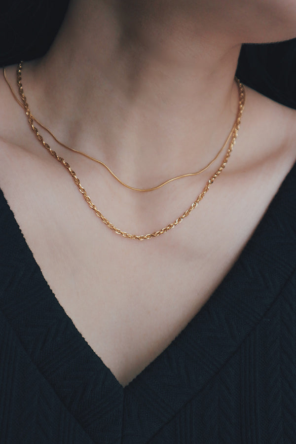A person wearing two layered gold chain necklaces, including a delicate snake chain and a finer textured chain, against a dark top.
