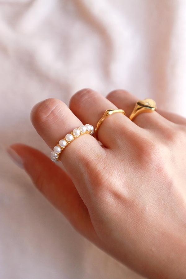 A hand wearing three stacked gold rings: a pearl band ring, an organic wavy gold ring, and a gold heart-shaped signet ring.