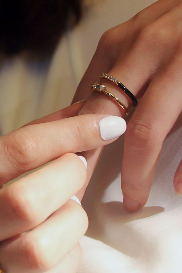 A close-up of a female hand with short, white manicured nails, adjusting two gold rings on her ring finger. One ring is a slender band, half black enamel and half adorned with clear cubic zirconia stones. The other ring on the same finger is a gold band with a small, light-colored bead and clear stones.