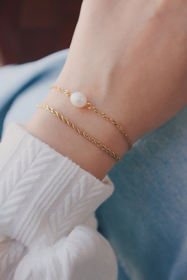 手腕上佩戴兩條疊戴金色手鍊:一條鑲有一顆白色珍珠的細緻鍊,和一條有紋理的麻花鍊。