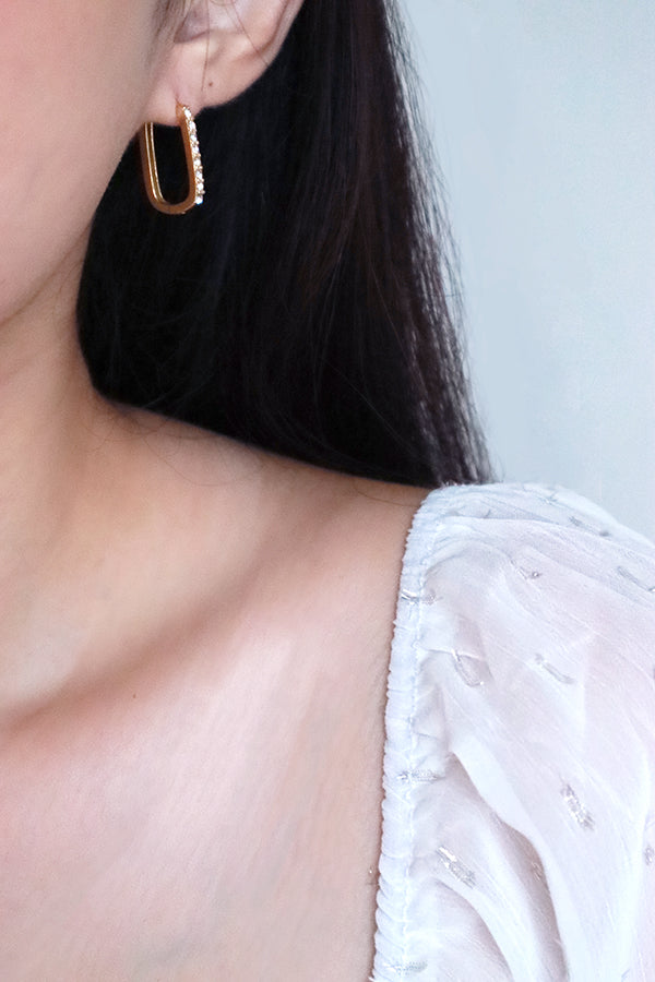 一位女性耳朵和肩部的側面輪廓,展示著一枚鑲嵌著一排閃亮透明寶石的長方形金色圈形耳環。這位女性留著長黑髮,穿著一件帶有細微銀色細節的白色薄紗上衣。