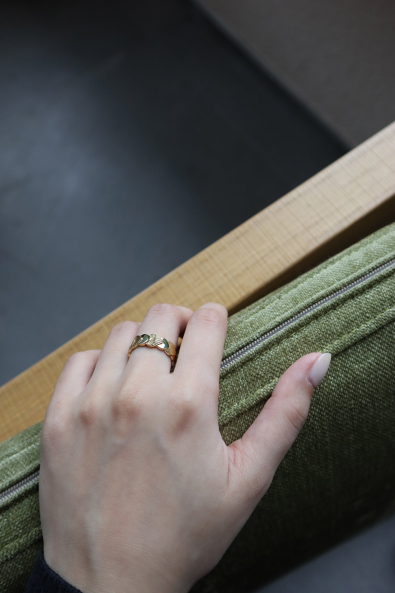 一隻女性手部特寫,輕放在綠色軟墊椅子的扶手上,無名指上戴著一枚帶有紋理扇形或貝殼設計的金戒指。背景模糊且黑暗。