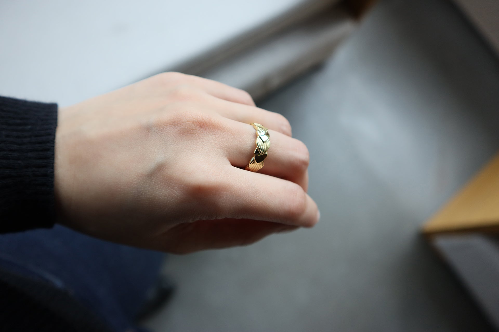 一隻留著淺色美甲的女性手部特寫,無名指上戴著一枚帶有紋理扇形或貝殼設計的金戒指。這位女性穿著黑色袖子,背景模糊且呈淺色。