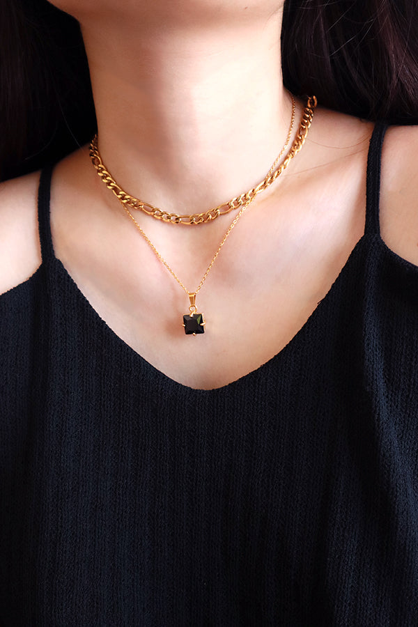A close-up of a woman's chest and neck, wearing two layered gold necklaces. The top necklace is a chunky chain, and the bottom is a delicate chain with a square-cut black rhinestone pendant. The woman is wearing a black camisole.