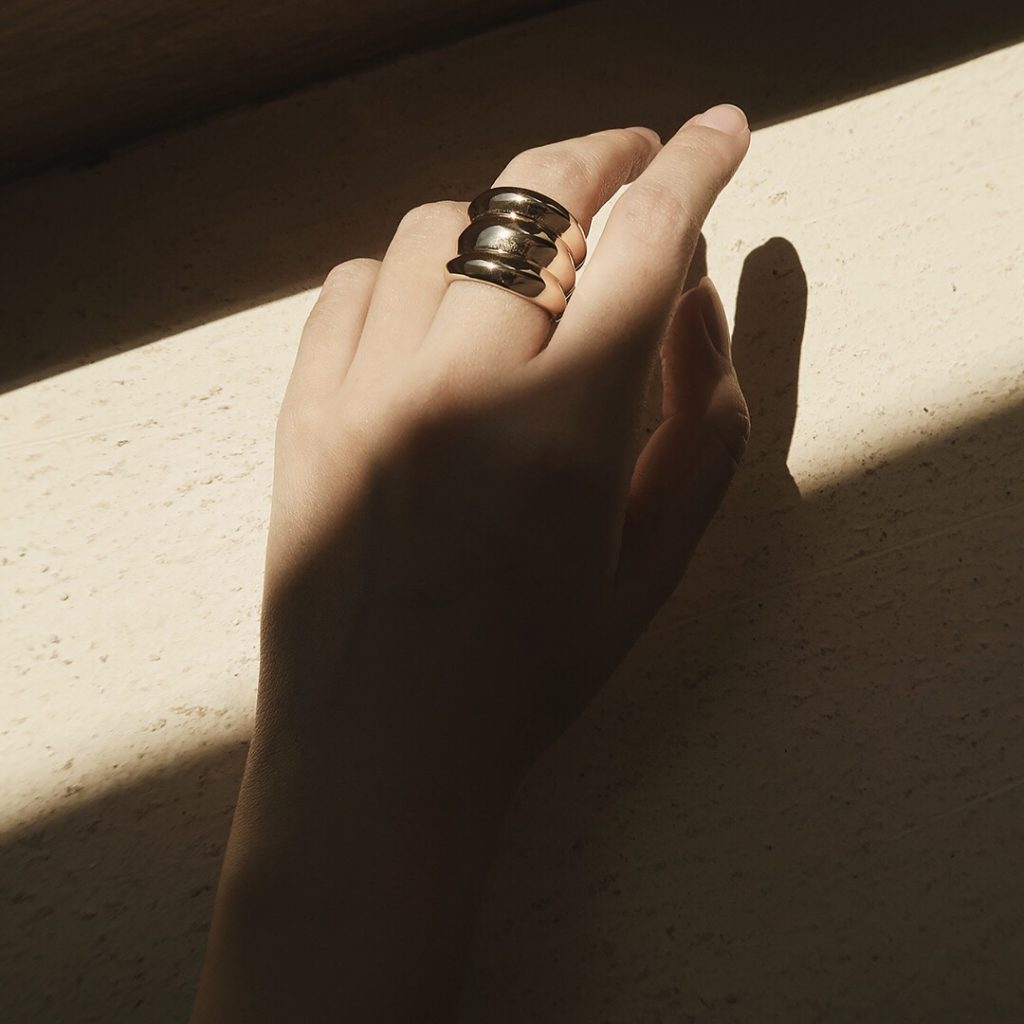 一隻女性手部特寫,無名指上戴著一枚顯眼的層疊金戒指。陽光在她的手上投下強烈的斜向陰影。背景是淺色紋理表面。