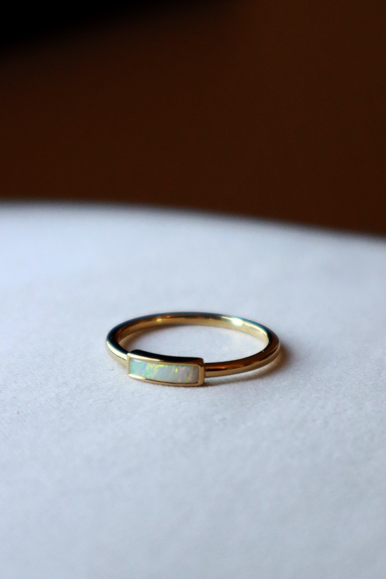 A close-up, eye-level shot of a minimalist gold ring with a rectangular, iridescent white opal inlay, resting on a light-colored surface with a blurred dark background.