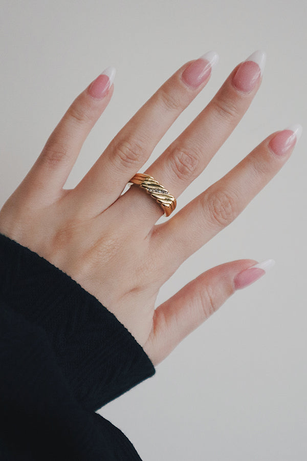 一隻留著淺色法式美甲的女性手,中指上戴著一枚厚重的金色戒指。戒指具有紋理條紋設計,並鑲嵌著一排斜向的小閃亮透明寶石。這位女性穿著一件黑色長袖上衣。