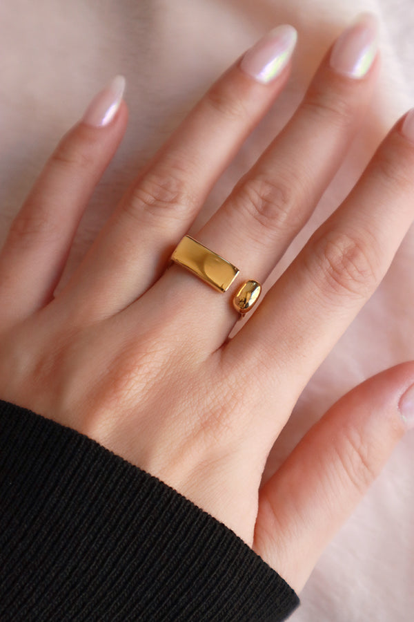 A person's hand wearing a fashionable 18k gold-plated open-ended ring with a rectangular bar and a small sphere, styled on a finger.
