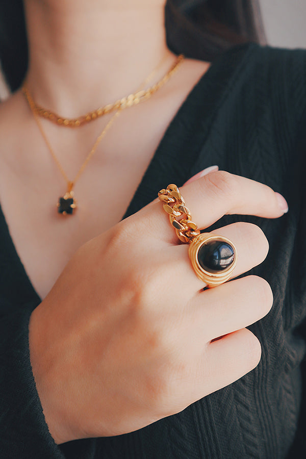 一隻膚色白皙的手,戴著一枚帶有大顆圓形黑色寶石的金色古巴鏈戒指。背景中,脖子上戴著一條帶有黑色吊飾的金色項鍊,穿著黑色上衣。