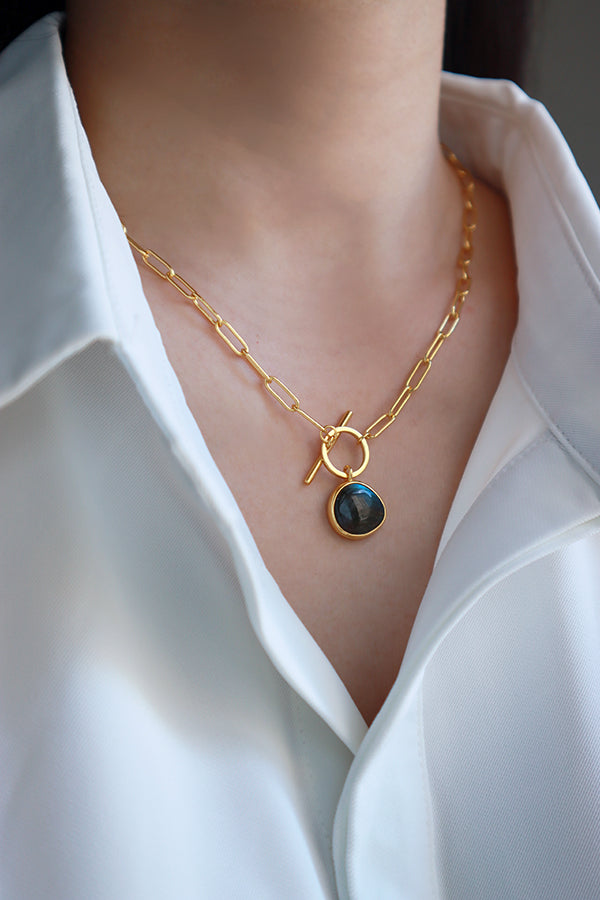 一位女性的頸部佩戴著一條時尚的金色迴紋針鍊條項鍊,項鍊中央飾有T字扣環,下方懸掛著一顆圓潤且閃爍著多重色彩光芒的拉長石寶石墜飾,與白色上衣完美搭配。